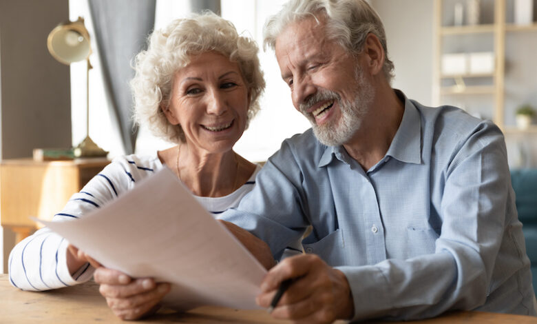 Smiling elderly 50s husband and wife sit at table in living room read contract agreement satisfied with terms and rules, happy old 60s couple customers or clients glad with pension health insurance