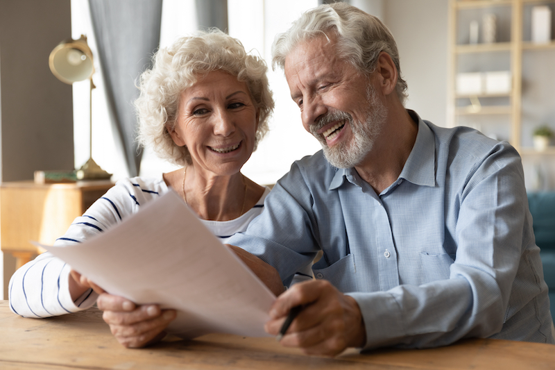 Smiling elderly 50s husband and wife sit at table in living room read contract agreement satisfied with terms and rules, happy old 60s couple customers or clients glad with pension health insurance