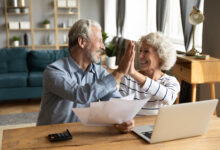 Overjoyed exited middle aged married couple giving high five, finishing doing domestic paperwork together at home. Euphoric happy older mature spouses celebrating successful investment or purchase.