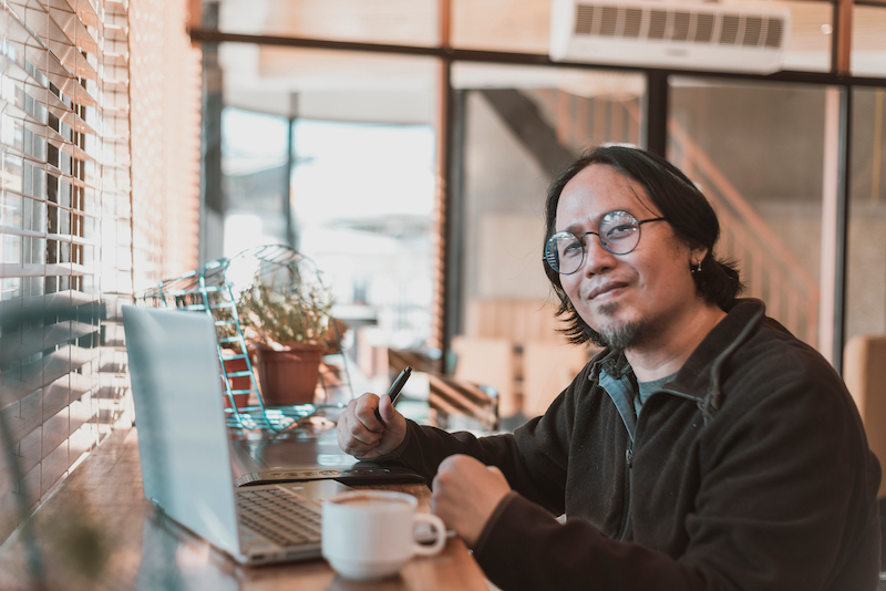 A middle aged asian graphic designer freelancer with a laptop and drawing tablet. Working remotely at a coffee shop or cafe.