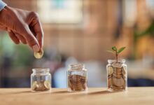 Close up of black woman hand adding money in coin in a jar. Girl hand holding coin adding money in glass jar of different sizes. Investment, savings and interest concept.