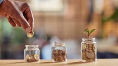 Close up of black woman hand adding money in coin in a jar. Girl hand holding coin adding money in glass jar of different sizes. Investment, savings and interest concept.