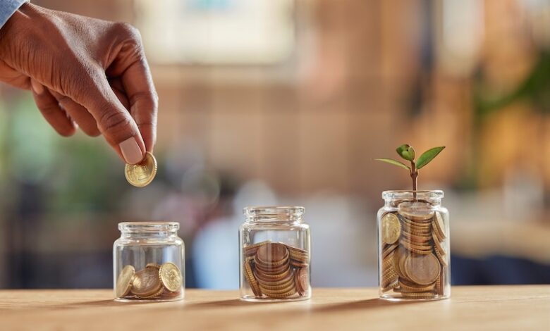 Close up of black woman hand adding money in coin in a jar. Girl hand holding coin adding money in glass jar of different sizes. Investment, savings and interest concept.