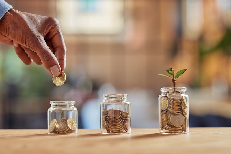 Close up of black woman hand adding money in coin in a jar. Girl hand holding coin adding money in glass jar of different sizes. Investment, savings and interest concept.